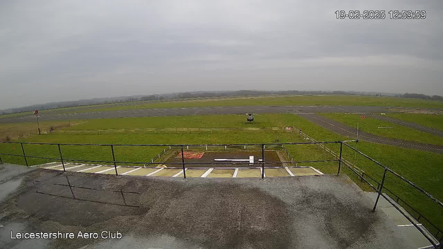 A view from a webcam showing a grassy area with a runway and some scattered markings on it. In the foreground, there is a railing and a flat surface. In the background, there is a helicopter on the ground, surrounded by a perimeter fence. The sky is clouded and gray, indicating an overcast day. The view suggests a rural setting, likely at an airfield or aero club.