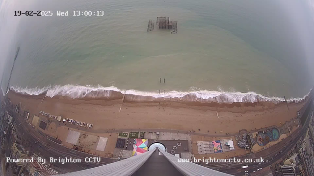 Aerial view of a beach scene with dark sand and shoreline along the ocean. Waves gently roll in from the water, creating white foam at the edge. Visible structures include a partially submerged pier in the water and various buildings along the beach, including a colorful amusement park area. The image captures a cloudy sky above, with a timestamp indicating the view is from February 19, 2025, at 1:00 PM.