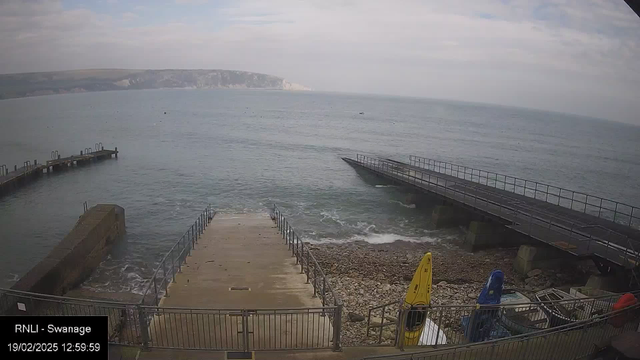 A seaside scene featuring a concrete ramp leading down to the water, with steps on either side. To the left, a short wooden pier juts out into the sea, and to the right, another pier extends further. The water is calm, with gentle waves, and small boats are visible in the distance. The background shows a rocky coastline under a cloudy sky. Various colored kayaks are stored on the right side near the pier.