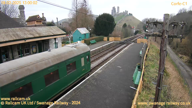 A green train carriage is parked at a railway station. The station features a stone building with a sloped roof and several large windows. In the background, there are hills with castle ruins visible at the top. The platform is lined with green benches and has a wooden fence on one side. There are also electric poles and a road beside the tracks. The sky is overcast, giving the scene a muted tone.