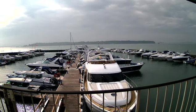 A view of a marina with several boats moored. The foreground features a wooden dock leading to a large white yacht, which is positioned among smaller boats of various colors and sizes, including blue and black. The scene is slightly overcast with gray clouds in the sky, and the water is calm, reflecting the boats and sky. In the distance, a faint outline of land can be seen across the water.