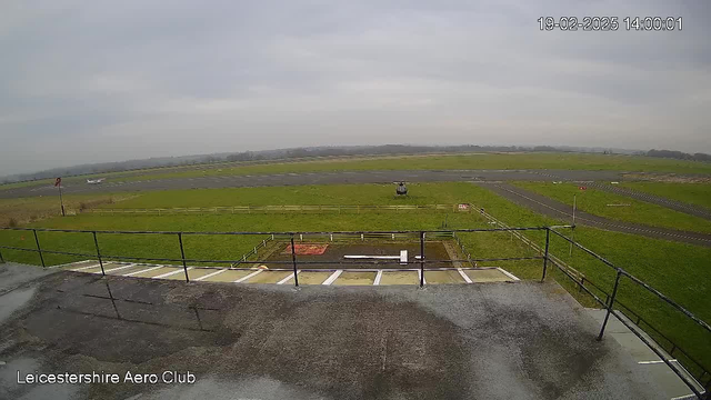 A cloudy sky is visible above a grassy field. In the foreground, there is a flat roof with a railing. A small aircraft is seen on the runway in the background, along with a helicopter parked nearby. Several signs and a fence are present along the edge of the grassy area. The location is identified as Leicestershire Aero Club, with a timestamp indicating the date and time of the image.