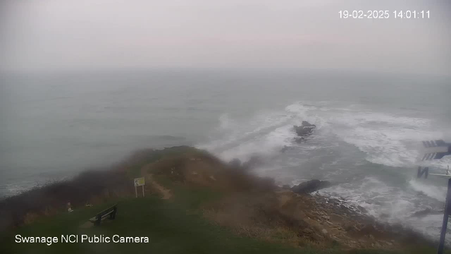 A cloudy sky with a grayish tint looms over a turbulent sea. Waves crash against rocky outcrops in the water, creating white foam. In the foreground, there is a patch of grassy land with a wooden bench, partially obscured by mist. A small sign is visible near the path leading to the edge. The scene conveys a sense of a stormy coastal area, with limited visibility due to the overcast conditions.