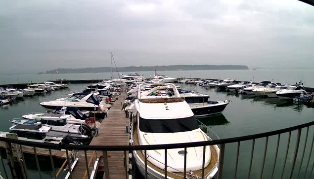 A cloudy marina scene featuring numerous boats docked at a wooden pier. The water is calm and reflects the overcast sky. In the foreground, a large white motorboat is anchored, surrounded by smaller boats of various sizes and colors. The marina extends into the background, with more boats visible and a distant shoreline. The atmosphere appears tranquil and serene.