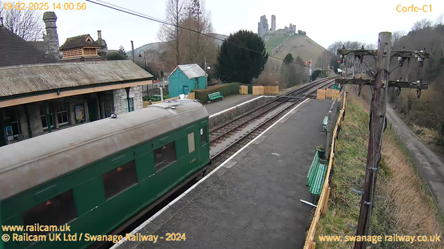 A green vintage train is partially visible on a platform at Corfe Castle railway station. In the background, the station's buildings and a small blue shed can be seen, along with wooden benches painted green. The railway tracks extend to the right of the image, leading towards a hill with ruins of a castle on top. The surroundings include bare trees and a gravel pathway. The sky is partly cloudy, indicating a cool day.