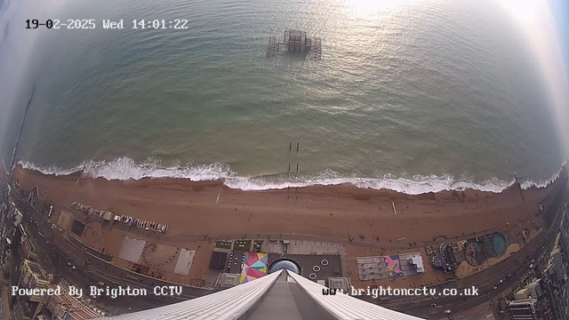 Aerial view of a beach with golden sand and gentle waves lapping at the shore. In the distance, a partially submerged pier is visible off the coast. There are various buildings, structures, and a colorful area with patterns near the shoreline. The sky is mostly clear with some clouds, and the image is timestamped at 14:01:22 on February 19, 2025. The bottom of the image shows part of a tall structure with a pointed tip. The image is marked by a logo indicating it is powered by Brighton CCTV.