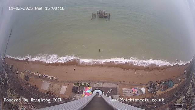 A high-angle view of a beach scene with gentle waves lapping against the shore. The sandy beach stretches along the bottom of the image, with various structures and an amusement park visible on the left side. In the distance, there is a weathered pier extending into the water. The sky is grey, cloudy, and there are a few distant boats on the horizon. The timestamp in the upper left corner indicates the date and time. The image is taken from a webcam, suggesting a surveillance or monitoring perspective.