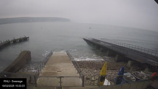 A foggy coastal scene featuring a calm sea with gentle waves. In the foreground, there is a concrete ramp leading down to the water, surrounded by a railing. To the left, a stone pier extends into the water. Two boats are moored, one yellow and one blue, on the right side near the shoreline. The background shows a distant cliff under a cloudy sky. The time and date are displayed in the bottom left corner.