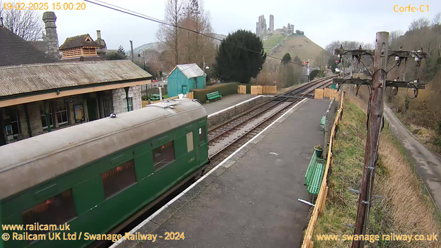 A green train carriage is positioned on the left side of the image, with its roof slightly visible. There is a railway platform with a textured concrete surface, featuring wooden benches painted green along the side. In the background, a hill with castle ruins can be seen, partially obscured by trees. To the right, a wooden utility pole with wires is visible, and in the distance, a wooded area and a narrow pathway can be seen. The sky is overcast, creating a subdued atmosphere.