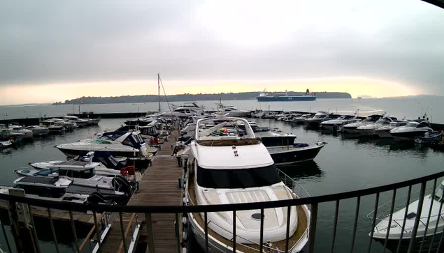 A marina filled with various boats docked in the water, with some larger ships visible in the background. The scene is taken from a high vantage point, showing a wooden walkway leading into the marina. The sky is overcast, with soft lighting creating a calm atmosphere. The water reflects the cloudy sky.