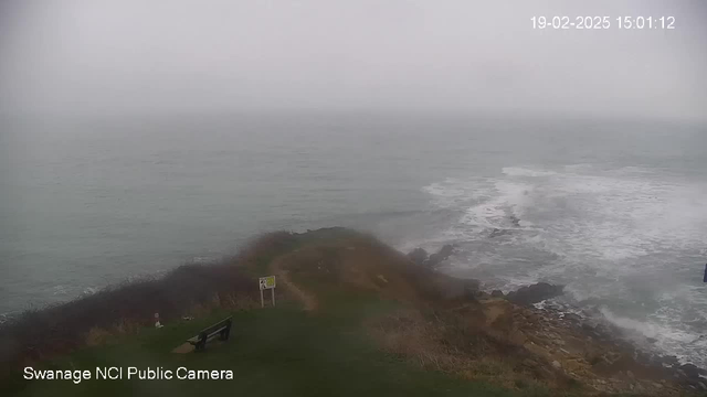 A coastal view during a foggy day. The sea is visible with gentle waves crashing against rocky formations. In the foreground, there is a grassy area with a bench and a warning sign. The sky is overcast, blending with the sea in shades of gray.