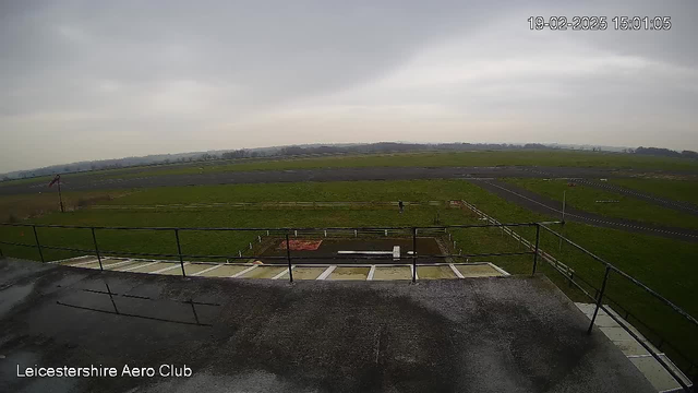 A view from a webcam overlooking a grassy field and an aerodrome. The scene is mostly cloudy, with a grey sky. In the foreground, there are rooftops and a railing, while the background features a large expanse of grass with some fencing. A few vehicles and markings are visible on the ground, along with a person walking in the distance. The timestamp at the top right indicates the date and time of the image.