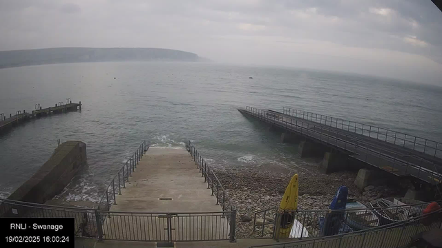 A view of a calm sea meeting the shore under a cloudy sky. In the foreground, a concrete ramp leads down to the water, bordered by a metal railing. To the left, there is a stone jetty extending into the water, while to the right, a wooden pier stretches out. Various boats are visible at the edge of the ramp, with one yellow kayak and others in different colors. The distant coastline is blurred by mist, giving a serene but overcast atmosphere.