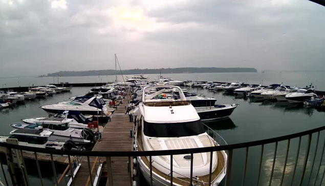 A view of a marina filled with numerous boats docked in calm waters under a cloudy sky. In the foreground, a larger white boat is prominently positioned, surrounded by various smaller boats. A wooden dock extends along the left side of the image, leading to more boats and a distant shoreline. The atmosphere appears tranquil with subdued lighting from the overcast sky.