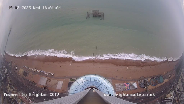 A bird's-eye view over a beach with sandy shore and waves lapping at the coast. In the distance, a derelict pier structure is partially submerged in the water. The foreground shows a circular glass structure, resembling a viewing platform, with a round roof. Along the beach, there are amusement rides, and a road lined with buildings. The scene is captured on a cloudy day, and the date and time are displayed in the top left corner of the image.