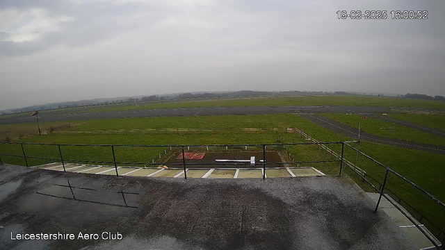 A wide view from a webcam shows a cloudy sky over an airfield. In the foreground, there is a flat rooftop with a railing, which has wet surfaces indicating recent rain. Below the rooftop, there is an airstrip lined with grass on either side. A small windsock can be seen to the left, indicating wind direction. The background features lush green fields and some trees in the distance, all under a gray overcast sky. The time stamp indicates the image was taken in the afternoon.