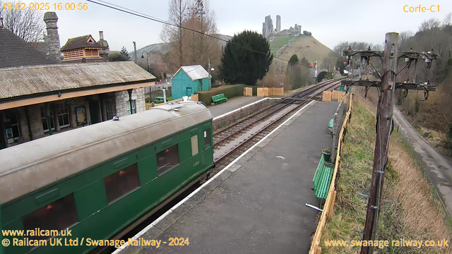 A green train carriage is positioned at a railway station with tracks extending into the distance. The station features a stone building with a sloped roof, and there is a blue shed nearby. Various green benches are visible along the platform. In the background, a hill rises with castle ruins at the top, partially covered by trees. The scene is set in a cloudy environment, and the station appears tranquil with no apparent activity.