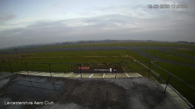 The image shows an open landscape viewed from a high position at the Leicestershire Aero Club. A flat, rooftop surface with a railing is visible in the foreground. Below, there is a green grassy area with a few visible pathways and a runway. The sky is overcast with soft clouds, and the scene appears to be slightly hazy. In the distance, there are fields that extend towards the horizon. A flagpole can be seen on the left side, suggesting there may be a flag at the top. The date and time displayed in the corner indicate it was taken on February 19, 2025, at 17:00:08.