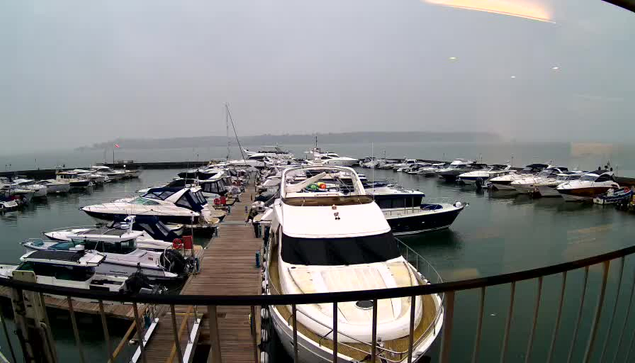 A view of a marina filled with various boats and yachts docked along a wooden pier. The water is calm and reflects the overcast sky, which appears hazy. Several boats are arranged closely together, with some larger boats at the forefront. The background shows a distant shoreline obscured by the mist. The scene conveys a serene yet gloomy atmosphere.