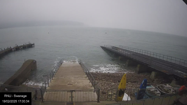 A foggy coastal scene shows a view of a calm sea. In the foreground, there are concrete stairs leading down to the water, flanked by metal railings. To the left, a wooden pier extends into the water. To the right, another pier is visible, which is wider and has a railing. Yellow and blue boats are stored on the stony shore, and the water's surface appears slightly rippled. The sky is overcast and gray, suggesting a misty atmosphere.