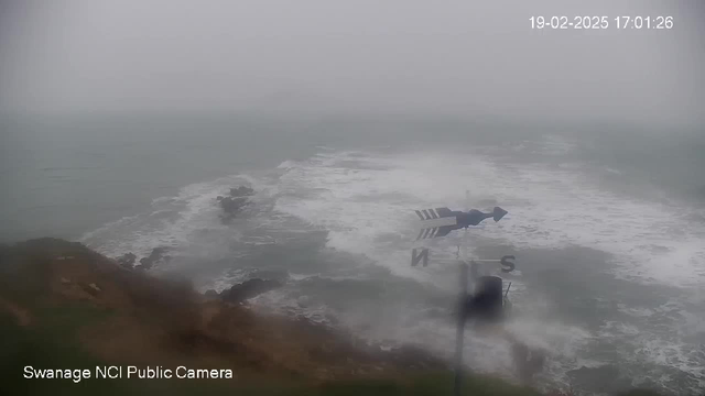 A hazy seascape captured by a webcam, showing rough waves crashing against rocky outcrops. The sky is overcast and gray, suggesting a stormy or misty day. In the foreground, a weather vane with directional markings is visible, turning slightly in the wind. The overall scene conveys a sense of tumultuous weather and coastal landscapes.