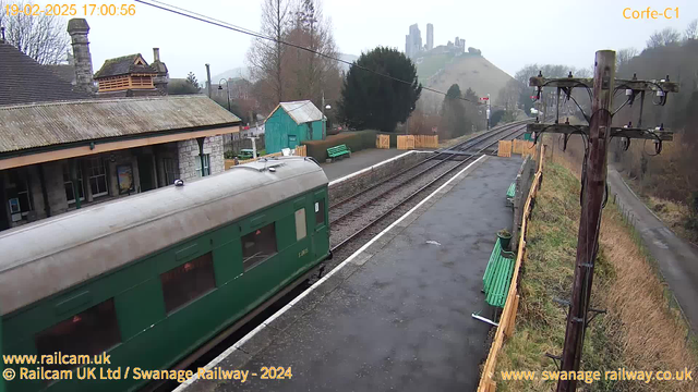 A narrow-gauge green train car is at a station platform, with the train's side visible. In the background, there are trees and a distant hill topped with ancient ruins. The platform has several benches, a green wooden shed, and wooden fencing. The sky is cloudy, creating a gray atmosphere. A section of railway track runs parallel to the platform.