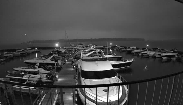 A view of a marina at night, featuring multiple boats docked in calm water. The scene is illuminated by faint lights from the boats and a distant pier. The sky appears overcast and dark, creating a moody atmosphere. In the foreground, a larger boat is visible, with a white hull and cabin, surrounded by smaller vessels. The reflections of the lights shimmer on the water's surface.