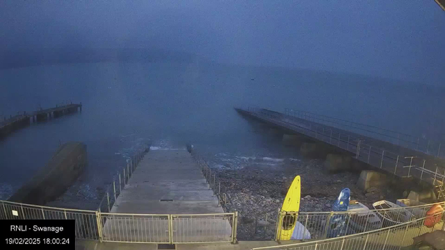 A dimly lit seaside scene shows a wooden jetty extending into calm water. Near the front, there is a sloped concrete ramp leading down to the water's edge, surrounded by a rocky shore. Two kayaks, one yellow and one blue, are parked on the right side, while another small boat is visible behind them. The sky is overcast, giving the image a muted blue tone. The time and date are noted in the corner, indicating it's early evening.