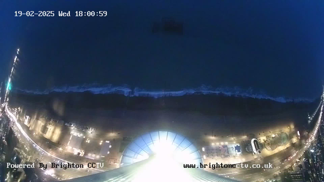 A nighttime webcam image showing a view of a beach and shoreline. The sea is visible with gentle waves lapping at the shore, against a dark background. A brightly lit area, possibly a pier or building, is at the bottom of the image, illuminating the surrounding area. Streetlights line the beach, casting a soft glow along the path. The date and time are displayed in the top left corner, and a watermark is present at the bottom.