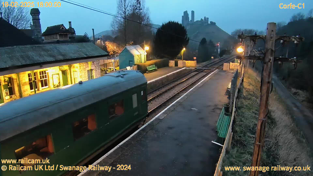 A green train carriage is positioned on a railway platform at dusk. The platform features several benches, some painted green, and is adorned with a couple of lampposts casting a warm glow. In the background, an old stone train station with lit windows adds to the scene, while a castle ruins sits atop a distant hill, partially obscured by trees. The sky is overcast and darkening as evening approaches. The railway track is visible, running parallel to the platform and leading into the distance.