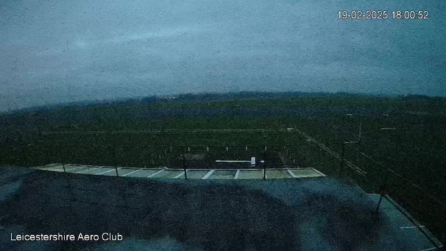 A dark, overcast scene of a field and runway from a webcam at Leicestershire Aero Club. The view shows a grassy area stretching into the distance, and a building rooftop in the foreground. There are faint outlines of structures or markers visible along the runway. The sky appears grey and cloudy, suggesting an evening time setting. The timestamp on the top right indicates the image was taken on February 19, 2025, at 18:00:52.