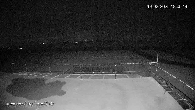 A dark scene from a webcam showing a large, empty tarmac area with a partial view of a low fence on the left side. The image is mostly black with faint lights visible in the distance, indicating a very low-light environment, possibly at night. The timestamp in the corner shows the date and time as 19-02-2025, 19:00:14.