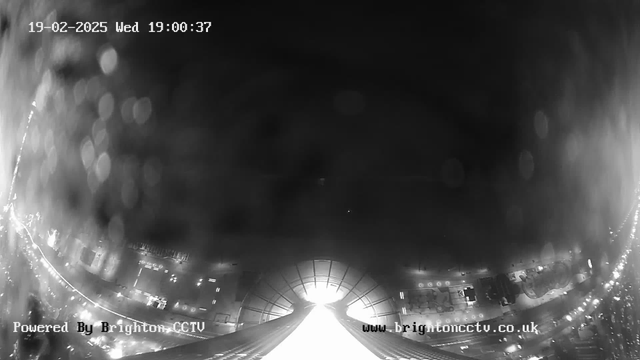 A black and white webcam image showing a city street at night. There is a bright light source at the center-bottom of the image, possibly indicating headlights or a reflective surface. The top half is darker with softer light spots that could be reflections or distant streetlights. The edges of the image appear slightly blurred. The time and date are displayed in the top left corner: "19-02-2025 Wed 19:00:37." The bottom left corner has the text "Powered By Brighton CCTV" and the website "www.brightoncctv.co.uk."