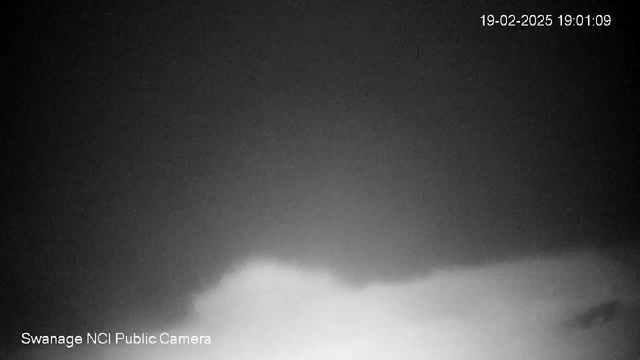 A dark image featuring a cloudy sky, with a gradient from darker shades at the top to lighter shades near the bottom. The time and date are displayed in the top right corner, showing "19-02-2025 19:01:09." The bottom left displays "Swanage NCI Public Camera." The overall atmosphere is somber and moody.