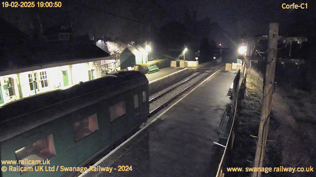 A dimly lit train station at night, with an old green train parked on the left side of the image. The platform is empty and features benches, with a small building in the background illuminated by soft light. There are decorative wooden fences and a lamppost casting light onto the scene, creating a quiet and serene atmosphere. Trees are visible in the background, partially obscured by darkness.