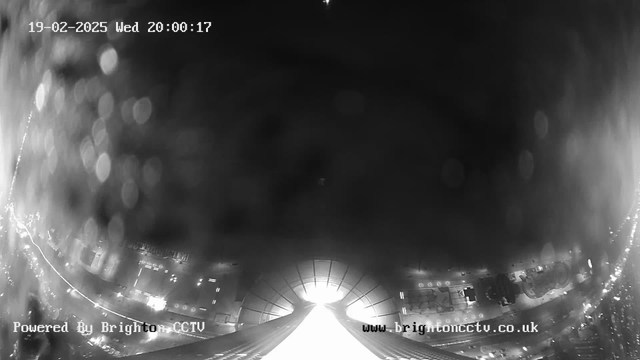 A black and white image from a webcam shows a view of a city at night. The bottom of the image features a bright light source, possibly a street or building, illuminating the area. Reflected light spots are visible throughout the image, creating a bokeh effect. The top of the image displays a faint star in the night sky, while the date and time are displayed in the upper left corner. The image has a grainy texture, suggesting low visibility conditions.