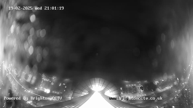 A nighttime webcam view featuring a dimly lit urban area. In the foreground, a bright pathway leads towards a tunnel-like structure. The scene is rainy, with droplets visible on the camera lens, creating a hazy effect. The timestamp in the upper left corner shows "19-02-2025 Wed 21:01:19." The bottom left corner displays the text "Powered By Brighton CCTV," while the right side of the image contains the website address "www.brightoncctv.co.uk."