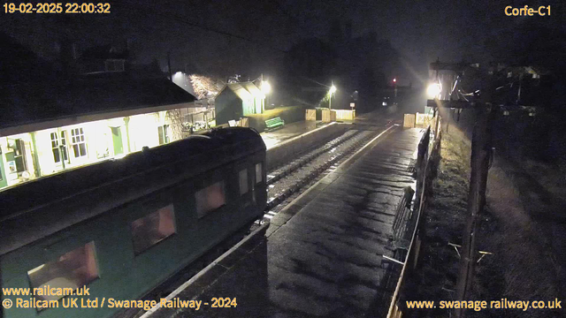 A dimly lit railway station scene at night. The foreground features a green train car with windows on the left. To the right, a platform stretches with a shiny surface reflecting light. A wooden fence and benches are visible further along the platform, illuminated by warm yellow light from nearby lamps. Shadows and deeper darkness surround the area, creating a contrast with the lit spaces. Trees can be faintly seen in the background, and there are utility poles with wires on the right side.