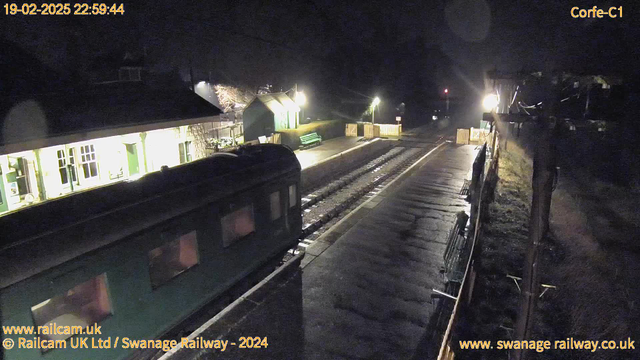 A nighttime view of a railway station. The foreground shows a green train car partially visible, with a shiny surface reflecting light. In the background, there is a small station building illuminated by warm lights, revealing windows and a door. A green bench is positioned beside the building. The platform is lined with rail tracks leading away, and wooden fencing is visible along the side. Streetlights cast a glow on the wet pavement, adding a soft brightness to the scene.