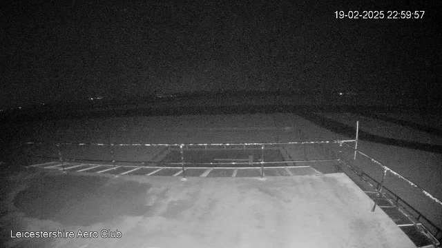 A dark scene captured by a webcam shows an empty outdoor area, possibly a runway or tarmac, illuminated by minimal ambient light. In the foreground, there is a railing along a flat surface, with faint markings indicating possible parking spaces. The background is mostly dark, with a few distant lights visible on the horizon, suggesting other buildings or vehicles are located further away. The time and date are displayed in the top right corner.