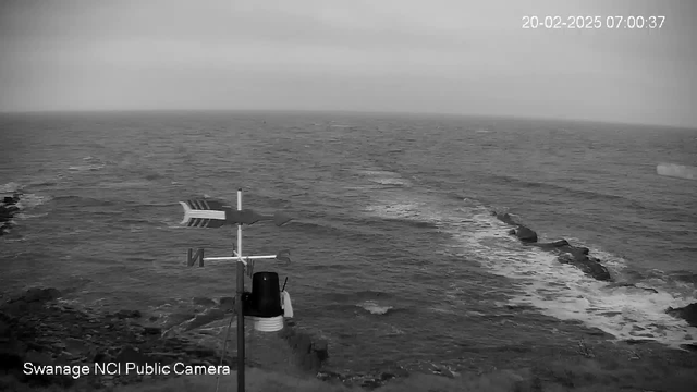 A black and white image showing a coastline with rough sea waves. In the foreground, there is a weather vane with direction indicators, primarily pointing north, and a black cylindrical structure next to it. The sea appears choppy, with a rocky shore visible on the left side of the image. The sky is overcast, suggesting a cloudy and possibly gloomy atmosphere. A timestamp indicates the image was taken on February 20, 2025, at 07:00:37.