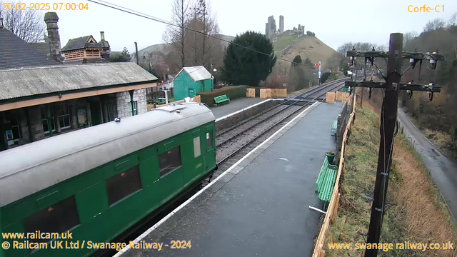 A green train car is parked at a railway station with a stone building visible in the background. The scene is set on a cloudy morning, and in the distance, old castle ruins are situated on a hill. The platform has a few green benches and a wooden fence lining the edge of the station area. The railway tracks run parallel to the platform, leading into the distance.