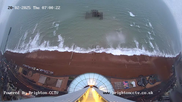 A bird's-eye view from a high vantage point looking down at a sandy beach beside the ocean. The image shows dark blue waves gently rolling in towards the shore, where the wet sand glistens. There is a pier structure partially submerged in the water, and closer to the bottom of the image, there's a walkway with various amenities visible. The sky is gray, indicating overcast weather, and the timestamp shows the date and time as February 20, 2025, at 7:00 AM.