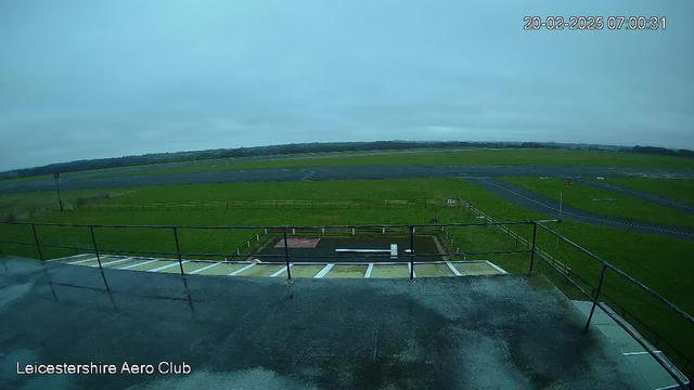 A cloudy sky fills the upper portion of the image. Below, an expansive grassy area leads to an airport runway, lined with a fence. The runway is dark and stretches horizontally across the landscape. Some markings and a red light tower are visible on the left side, while a small landing area is seen near the bottom of the frame, featuring a red square and a white strip. The scene appears wet, suggesting recent rain. The time and date are displayed in the corner.