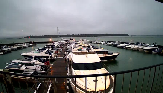 A view of a marina filled with numerous boats and yachts, some docked along a wooden pier. The scene is overcast, with a grey sky and calm water in the background. The boats are various sizes and colors, with one prominent white yacht at the center. Some boats have colorful covers and equipment visible on them.