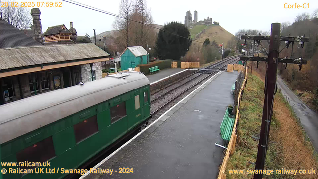 A green train car is positioned partially in the frame on the left side, with its door and windows visible. The scene shows a railway station with a covered platform. In the background, there is a hill with ruins, possibly castle remnants, at the top. The sky is overcast, and the ground appears wet, suggesting recent rain. There are benches along the platform, some with green paint. On the right, a wooden fence marks the boundary of the station, and a telephone pole is visible with wires running along the top of the image.