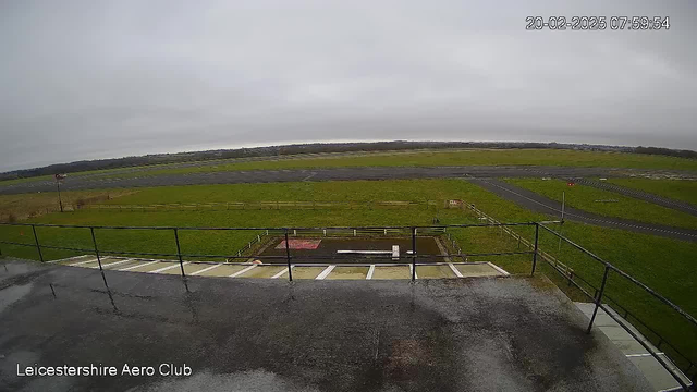 A view from a webcam situated at Leicestershire Aero Club, showing an overcast sky with clouds. In the foreground, there is a railing and a section of a flat roof. Below, a grassy area is visible along with a runway that stretches into the distance. The runway appears wet, likely from recent rain. There are also some markings and a sign near the edge of the runway, with green fields surrounding it. The scene conveys a quiet and rural setting, typical of an airport.