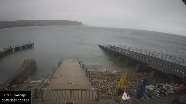 A cloudy view of a coastal area, with a mix of light and dark gray skies. In the foreground, a concrete ramp leads down to the water, bordered by a metal railing. To the left, there is a rocky area and a small harbor with several posts extending into the water. The right side shows a pier with a wide walkway extending into the sea. There are yellow, blue, and red kayaks stored on the shore, and the water is calm with a few small ripples. In the distance, a green and rocky coastline can be seen, and the overall scene appears tranquil and overcast.