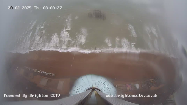 A high-angle view of a beach and ocean taken from a webcam. The image shows the waves crashing onto a sandy shore, with a pier visible in the water. There is a clear sky and some clouds, indicating overcast weather. Various structures and a few vehicles are scattered along the beach, partially obscured by the angle of the shot. The date and time in the corner indicate it's early morning on February 20, 2025. The bottom edge of the image displays text that identifies the source as Brighton CCTV.