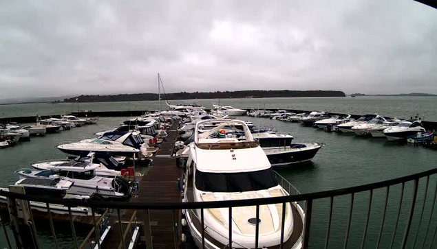 A marina scene under a cloudy sky, filled with numerous motorboats and yachts docked at a wooden pier. The water appears calm with a slight ripple, and some boats are tied up while others are anchored. In the background, the shoreline is visible, lined with trees and hills. The atmosphere is overcast, suggesting a possibility of rain.